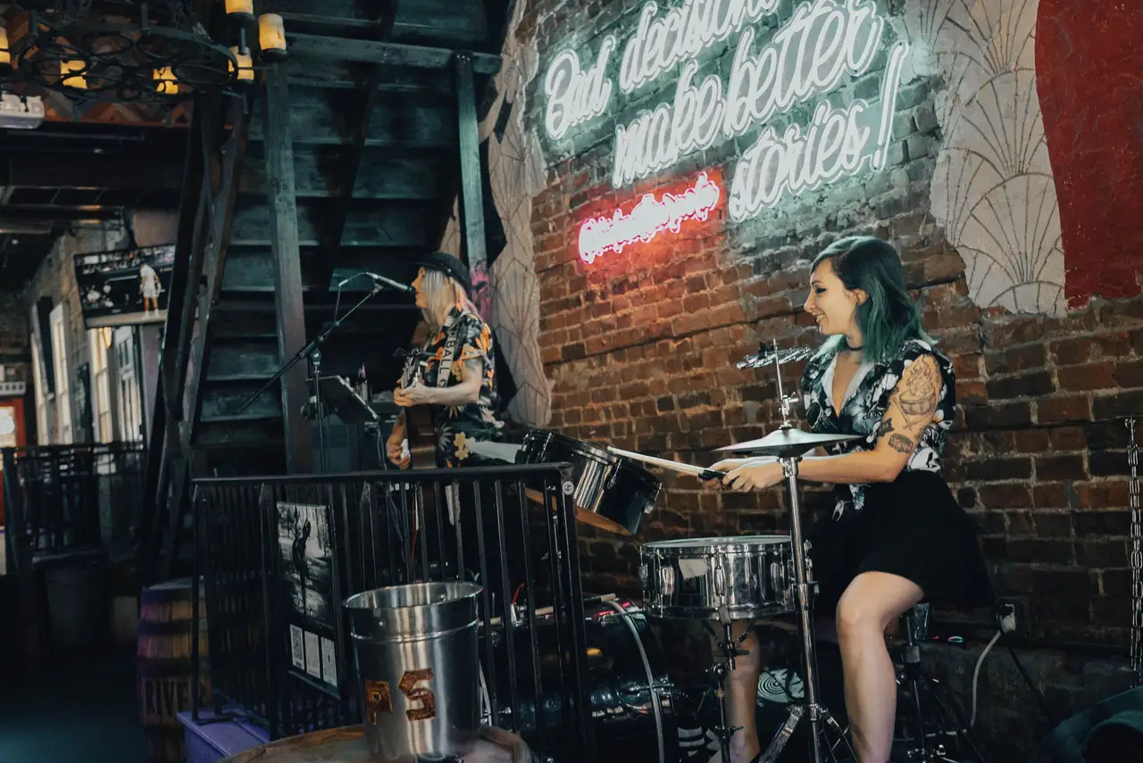 A guitarist and a drummer playing on The Hideout’s stage under a neon sign on the brick wall
