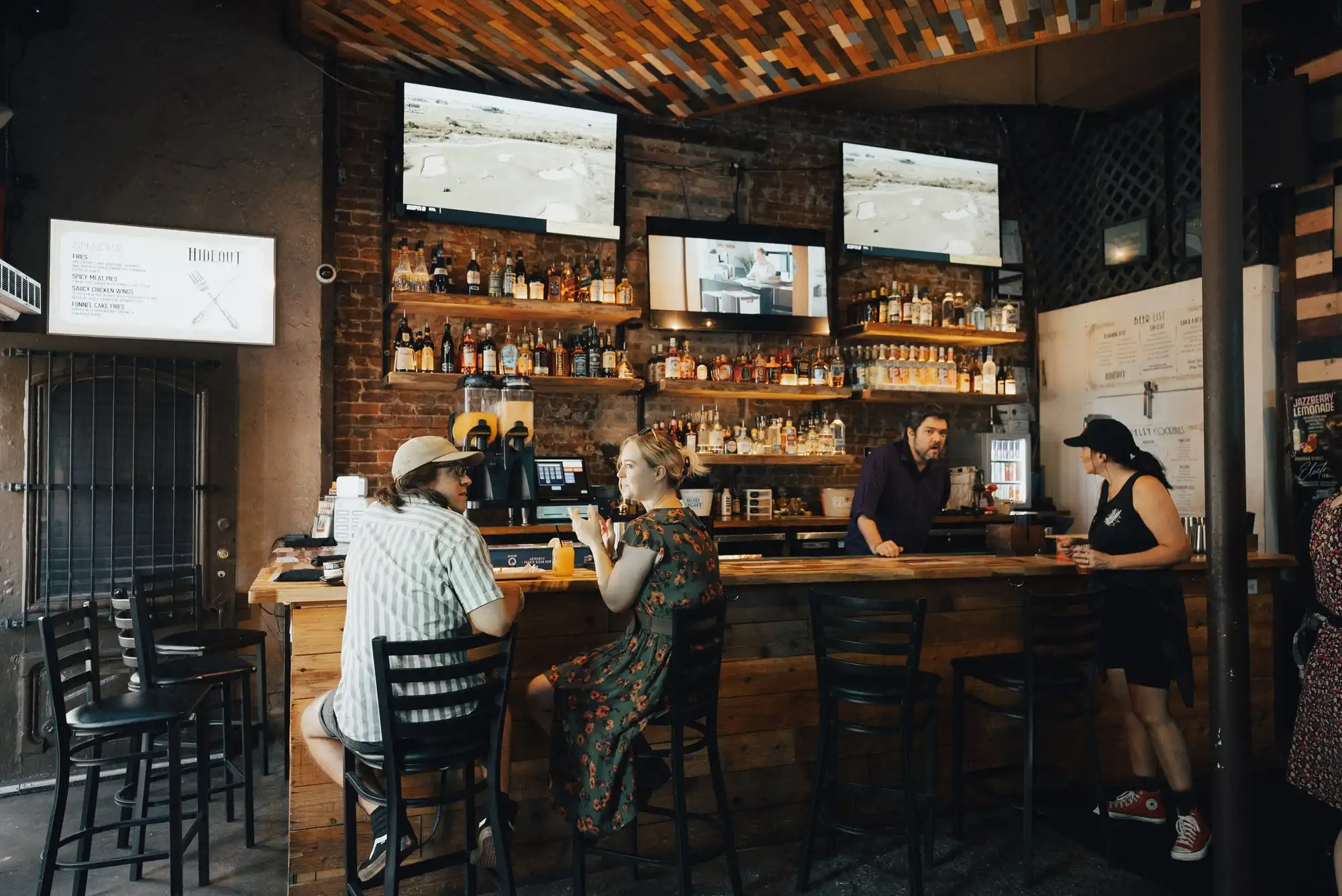 The front bar at The Hideout: guests on stools at a long wooden bar, with TVs and backlit bottle shelves on an exposed-brick wall