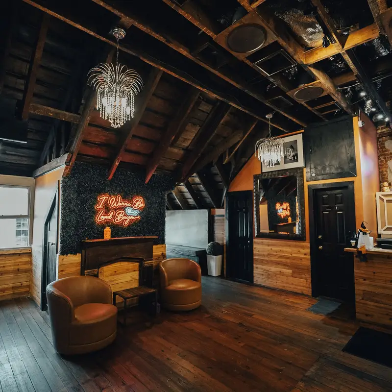 The Speakeasy Room under a crystal chandelier: lounge chairs, a neon sign, wood-paneled walls and a small bar in the corner